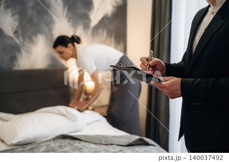 Hotel manager observing housekeeping staff while inspecting a neatly made bed in a modern hotel room with stylish decor and soft lighting 140037492