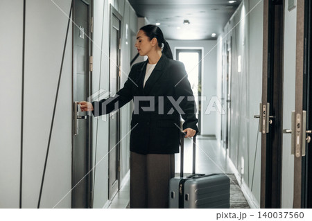 Woman with suitcase unlocking hotel room door in a modern corridor, featuring gray walls and contemporary design elements, showcasing a travel scenario Woman with suitcase unlocking hotel room door in a modern corridor, featuring gray walls and contemporary design elements, showcasing a travel scenario 140037560