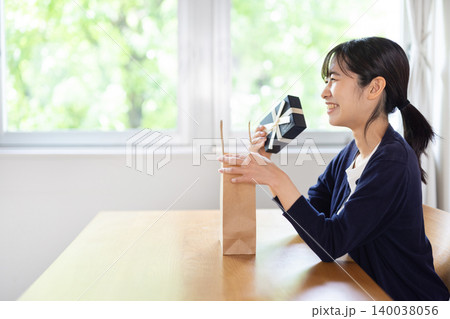 明るい室内で紙袋からプレゼント箱を取り出して微笑む日本人の若い女性 明るい室内で紙袋からプレゼント箱を取り出して微笑む日本人の若い女性 140038056