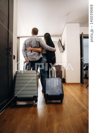 Couple standing together with luggage in a hotel room, embracing each other while facing the interior, wooden floor and modern furnishings visible in the background Couple standing together with luggage in a hotel room, embracing each other while facing the interior, wooden floor and modern furnishings visible in the background 140039069