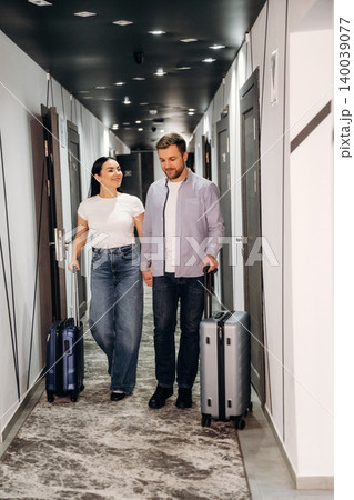 Couple walking together in a hotel corridor, carrying suitcases, with modern doors and stylish lighting visible along the hallway Couple walking together in a hotel corridor, carrying suitcases, with modern doors and stylish lighting visible along the hallway 140039077