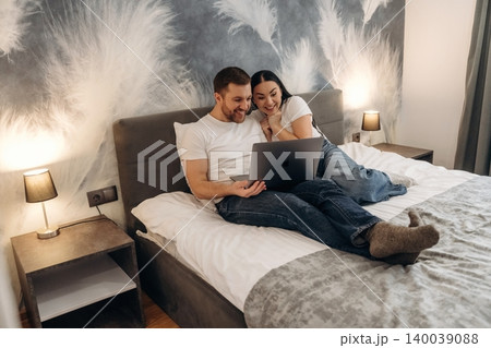 Couple sitting on a bed in a hotel room, using a laptop together, with decorative wall and bedside lamps providing ambient lighting Couple sitting on a bed in a hotel room, using a laptop together, with decorative wall and bedside lamps providing ambient lighting 140039088