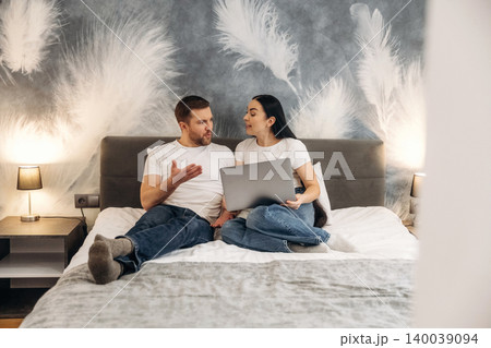 Couple sitting on bed in hotel room, discussing while using laptop, with decorative feather wall art and bedside lamps illuminating the cozy space Couple sitting on bed in hotel room, discussing while using laptop, with decorative feather wall art and bedside lamps illuminating the cozy space 140039094