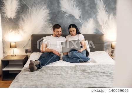 Couple sitting on a bed in a hotel room, using a laptop together, with decorative wall and bedside lamps creating a cozy atmosphere Couple sitting on a bed in a hotel room, using a laptop together, with decorative wall and bedside lamps creating a cozy atmosphere 140039095