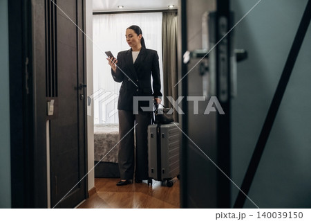 Businesswoman in black suit holding smartphone and suitcase stands in hotel room doorway, modern decor and bed visible in the background 140039150