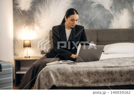 Businesswoman in black suit working on laptop while sitting on hotel bed with stylish decor and suitcase nearby, showcasing modern hotel interior design Businesswoman in black suit working on laptop while sitting on hotel bed with stylish decor and suitcase nearby, showcasing modern hotel interior design 140039162