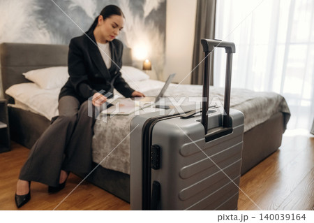 Businesswoman seated on hotel bed working on laptop with documents, gray suitcase positioned in foreground, modern interior design and natural light from window Businesswoman seated on hotel bed working on laptop with documents, gray suitcase positioned in foreground, modern interior design and natural light from window 140039164