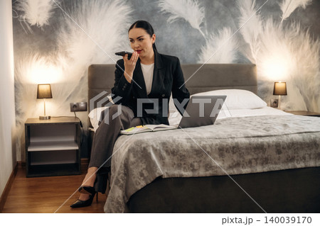 Businesswoman in black suit sitting on bed in hotel room, using smartphone while laptop and notebook are placed on bed with decorative wall in background 140039170