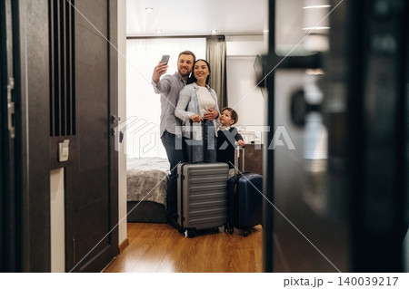 Family of three taking a selfie inside a hotel room, with luggage positioned near the entrance and a bed visible in the background Family of three taking a selfie inside a hotel room, with luggage positioned near the entrance and a bed visible in the background 140039217