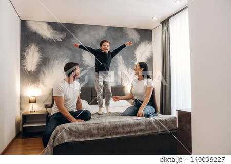 Family enjoying time together in a modern hotel room with a child jumping on the bed and parents sitting on the edge, decorated with feather-patterned wall art Family enjoying time together in a modern hotel room with a child jumping on the bed and parents sitting on the edge, decorated with feather-patterned wall art 140039227