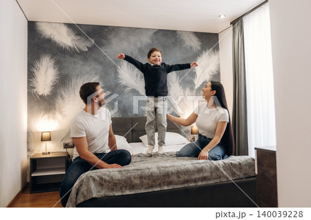 Family enjoying time together in a hotel room with a child jumping on the bed, modern decor featuring feather-patterned wall and soft lighting Family enjoying time together in a hotel room with a child jumping on the bed, modern decor featuring feather-patterned wall and soft lighting 140039228