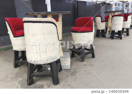 Rustic outdoor cafe furniture made from wooden barrels with red velvet seats, creating a cozy vintage street dining atmosphere 140039304