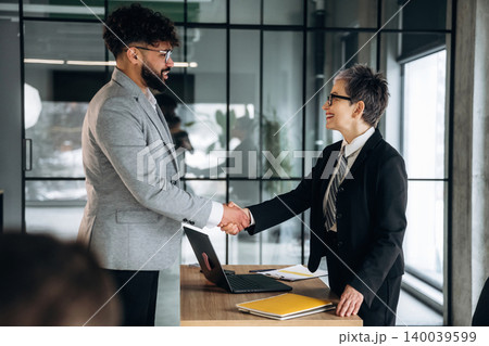 Business professionals shaking hands in a modern office setting, with a laptop and documents on the table, large windows providing natural light Business professionals shaking hands in a modern office setting, with a laptop and documents on the table, large windows providing natural light 140039599