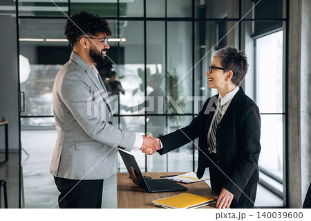Business professionals shaking hands in a modern office setting, with a laptop and documents on a wooden table, large windows providing natural light Business professionals shaking hands in a modern office setting, with a laptop and documents on a wooden table, large windows providing natural light 140039600