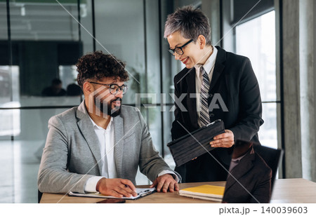 Business meeting in modern office with two professionals discussing project details at a wooden table, digital tablet and documents visible in a bright workspace 140039603