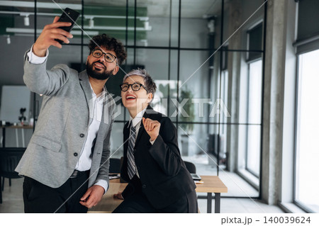 Two business professionals taking a selfie in a modern office setting with large windows and a wooden table, showcasing a collaborative work environment Two business professionals taking a selfie in a modern office setting with large windows and a wooden table, showcasing a collaborative work environment 140039624