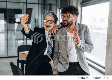 Two business professionals taking a selfie in a modern office environment, showcasing a stylish interior with large windows and contemporary furniture 140039629