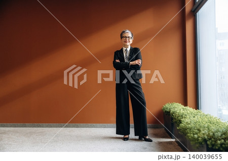 Businesswoman in black suit stands confidently with arms crossed in modern office space featuring a warm brown wall and green plants along the floor Businesswoman in black suit stands confidently with arms crossed in modern office space featuring a warm brown wall and green plants along the floor 140039655