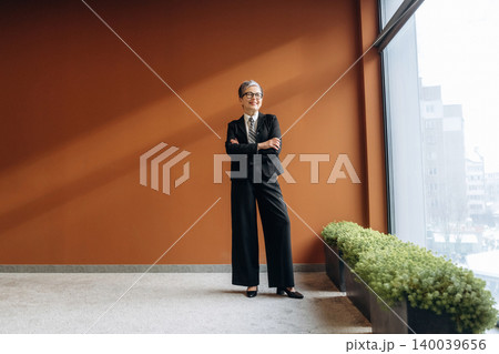 Businesswoman in black suit stands confidently with arms crossed in modern office space featuring large windows and green plants against an orange wall Businesswoman in black suit stands confidently with arms crossed in modern office space featuring large windows and green plants against an orange wall 140039656