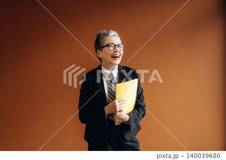 Businesswoman with short gray hair wearing a black suit and glasses holds a yellow folder while smiling in front of a brown wall in a modern office setting 140039680