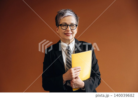 Senior businesswoman with short gray hair wearing glasses and a black suit holds a yellow notebook while standing against a brown background in an office setting 140039682