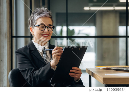 Businesswoman in black suit and glasses smiles while holding clipboard and pen, seated at desk in modern office with large windows and plants in the background Businesswoman in black suit and glasses smiles while holding clipboard and pen, seated at desk in modern office with large windows and plants in the background 140039684
