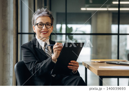 Mature Caucasian businesswoman in black suit holding clipboard and pen, smiling while seated at a modern office desk with documents and plants in the background 140039693