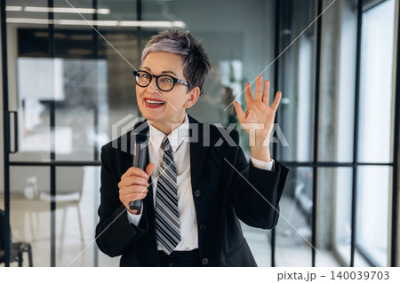 Businesswoman with short gray hair in black suit and tie holds microphone while speaking in modern office with glass walls and contemporary furniture Businesswoman with short gray hair in black suit and tie holds microphone while speaking in modern office with glass walls and contemporary furniture 140039703