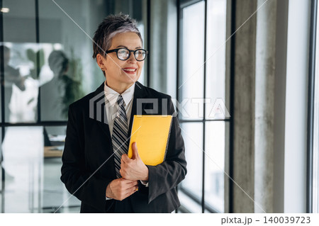 Businesswoman with short gray hair wearing a black suit and glasses holds a yellow notebook while standing in a modern office corridor with large windows Businesswoman with short gray hair wearing a black suit and glasses holds a yellow notebook while standing in a modern office corridor with large windows 140039723