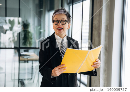 Businesswoman with short gray hair wearing a black suit and glasses holds yellow folders while standing in a modern office with large windows and greenery 140039727