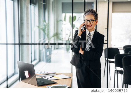 Businesswoman in black suit talking on smartphone while standing at desk with laptop and documents in modern office with glass walls and plants Businesswoman in black suit talking on smartphone while standing at desk with laptop and documents in modern office with glass walls and plants 140039753