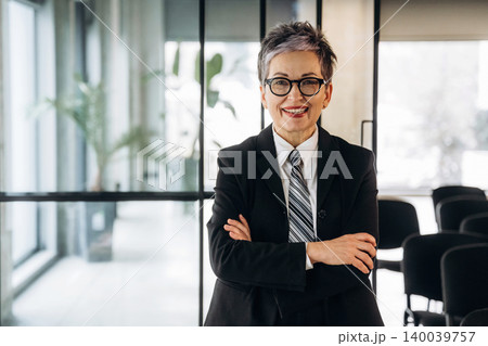 Businesswoman with short gray hair wearing a black suit and glasses stands confidently with arms crossed in a modern office space with glass walls and chairs 140039757