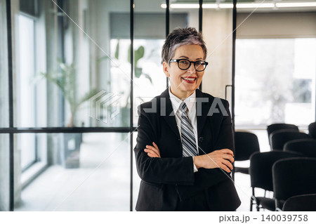 Businesswoman with short gray hair wearing glasses and a black suit stands confidently with arms crossed in a modern office space with glass walls and chairs Businesswoman with short gray hair wearing glasses and a black suit stands confidently with arms crossed in a modern office space with glass walls and chairs 140039758