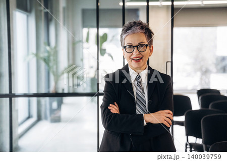 Businesswoman with short gray hair wearing a black suit and tie stands confidently with arms crossed in a modern office space with glass walls and chairs Businesswoman with short gray hair wearing a black suit and tie stands confidently with arms crossed in a modern office space with glass walls and chairs 140039759