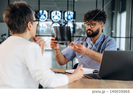 Male healthcare professional in blue shirt examines X-ray film while discussing results with female patient in modern medical office with laptop and medical images in background 140039868