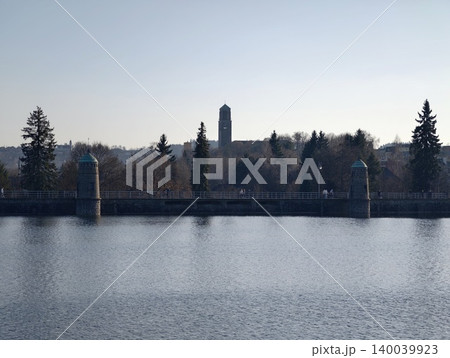 Calm reservoir with distant towers. Serene waterbody featuring industrial silhouettes and reflections Calm reservoir with distant towers. Serene waterbody featuring industrial silhouettes and reflections 140039923