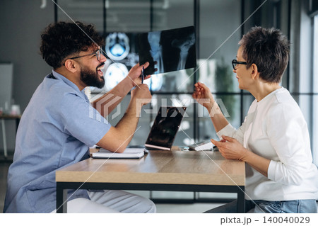 Male healthcare professional in blue scrubs discusses X-ray results with female patient in modern clinic setting, laptop and medical images visible on table 140040029