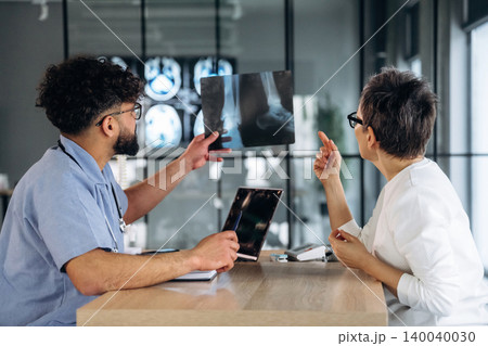 Male healthcare professional in blue scrubs discusses X-ray results with female doctor in white coat, modern medical office with large windows and equipment visible Male healthcare professional in blue scrubs discusses X-ray results with female doctor in white coat, modern medical office with large windows and equipment visible 140040030