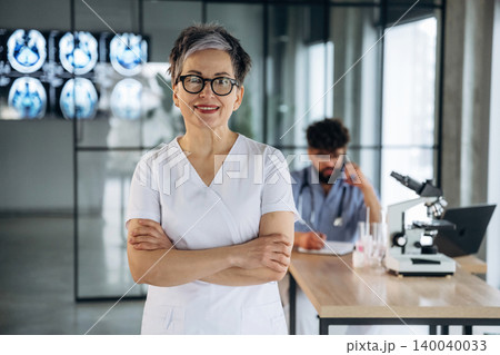Female healthcare professional with glasses stands confidently in a modern medical office, with a male colleague working at a desk and medical equipment visible in the background Female healthcare professional with glasses stands confidently in a modern medical office, with a male colleague working at a desk and medical equipment visible in the background 140040033