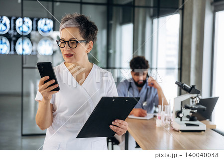 Female healthcare professional in white scrubs using smartphone while holding clipboard, with male colleague studying medical samples in modern laboratory setting Female healthcare professional in white scrubs using smartphone while holding clipboard, with male colleague studying medical samples in modern laboratory setting 140040038
