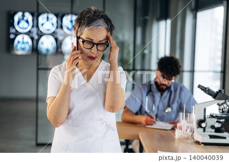 Female healthcare professional in white scrubs holds her head while talking on the phone, with a male colleague and medical equipment in a modern clinic setting Female healthcare professional in white scrubs holds her head while talking on the phone, with a male colleague and medical equipment in a modern clinic setting 140040039