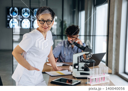 Female healthcare professional in white scrubs smiles confidently while standing beside a microscope and test tubes in a modern medical office with a colleague in the background Female healthcare professional in white scrubs smiles confidently while standing beside a microscope and test tubes in a modern medical office with a colleague in the background 140040056