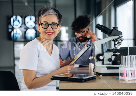 Female healthcare professional in white scrubs smiles while holding a clipboard, with a microscope and laboratory equipment visible in a modern medical office Female healthcare professional in white scrubs smiles while holding a clipboard, with a microscope and laboratory equipment visible in a modern medical office 140040059