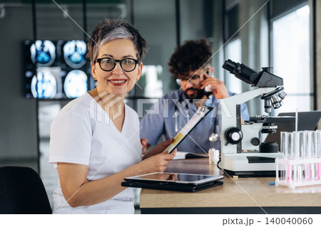 Female healthcare professional with short gray hair and glasses holds a clipboard while sitting at a laboratory table with a microscope and test tubes in a modern medical facility Female healthcare professional with short gray hair and glasses holds a clipboard while sitting at a laboratory table with a microscope and test tubes in a modern medical facility 140040060