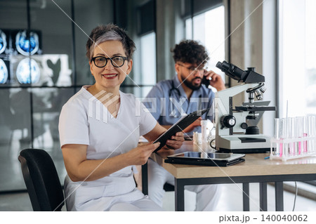 Female healthcare professional in white scrubs holding a clipboard, seated at a laboratory table with a microscope and test tubes, male colleague in background on phone Female healthcare professional in white scrubs holding a clipboard, seated at a laboratory table with a microscope and test tubes, male colleague in background on phone 140040062