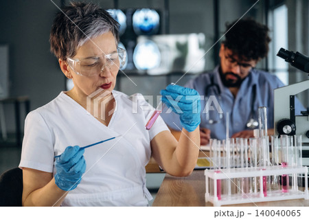 Female healthcare professional in laboratory coat examines test tube with pink liquid while male colleague works at microscope in modern laboratory setting Female healthcare professional in laboratory coat examines test tube with pink liquid while male colleague works at microscope in modern laboratory setting 140040065