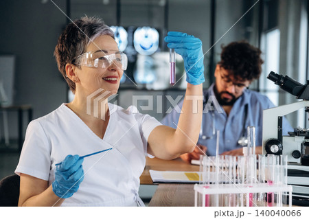 Female laboratory technician in white coat examines test tube with pink liquid while male colleague works at desk with microscope and documents in modern healthcare facility Female laboratory technician in white coat examines test tube with pink liquid while male colleague works at desk with microscope and documents in modern healthcare facility 140040066