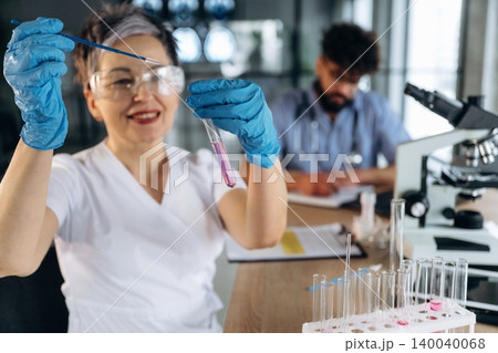 Female laboratory technician in white coat holds test tube with pink liquid, wearing blue gloves, with male colleague and microscope in modern lab setting Female laboratory technician in white coat holds test tube with pink liquid, wearing blue gloves, with male colleague and microscope in modern lab setting 140040068