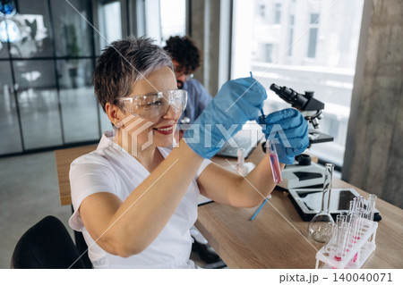 Female scientist in laboratory wearing safety goggles and gloves, holding test tube with colorful liquid while working at a wooden desk with laboratory equipment in the background 140040071