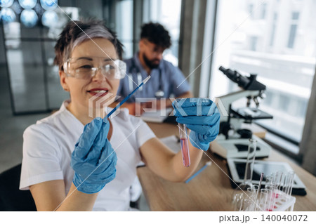 Female laboratory technician in white coat holds test tube with pink liquid and pipette, with microscope and male colleague in background inside modern lab 140040072